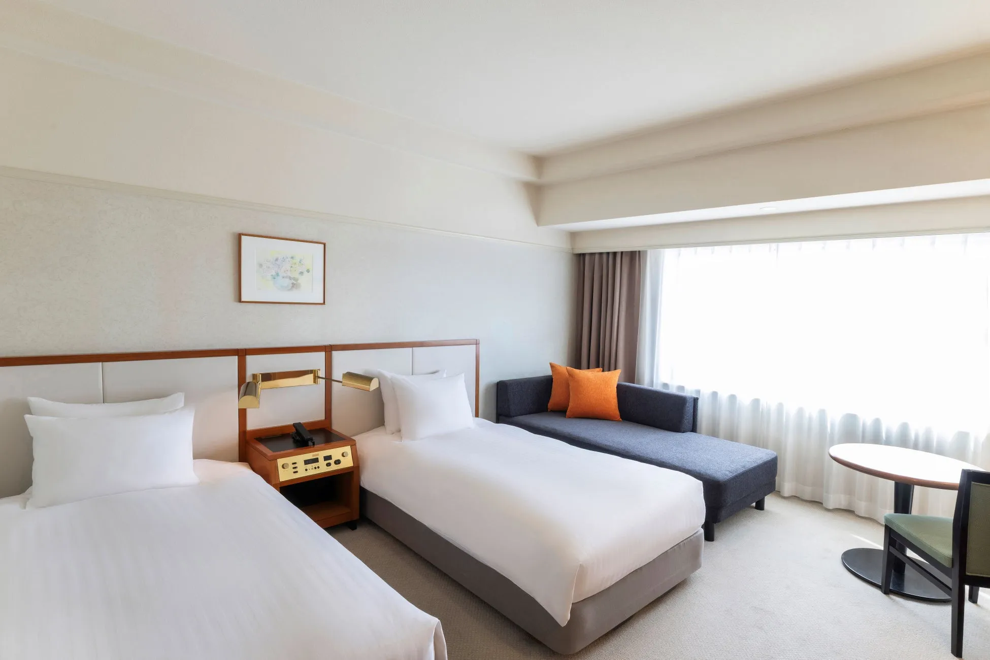 ホテルの客室の写真です。ツインベッドが2つあり、ソファ、丸テーブル、椅子が置かれています。窓からは自然光が差し込み、明るい雰囲気です。