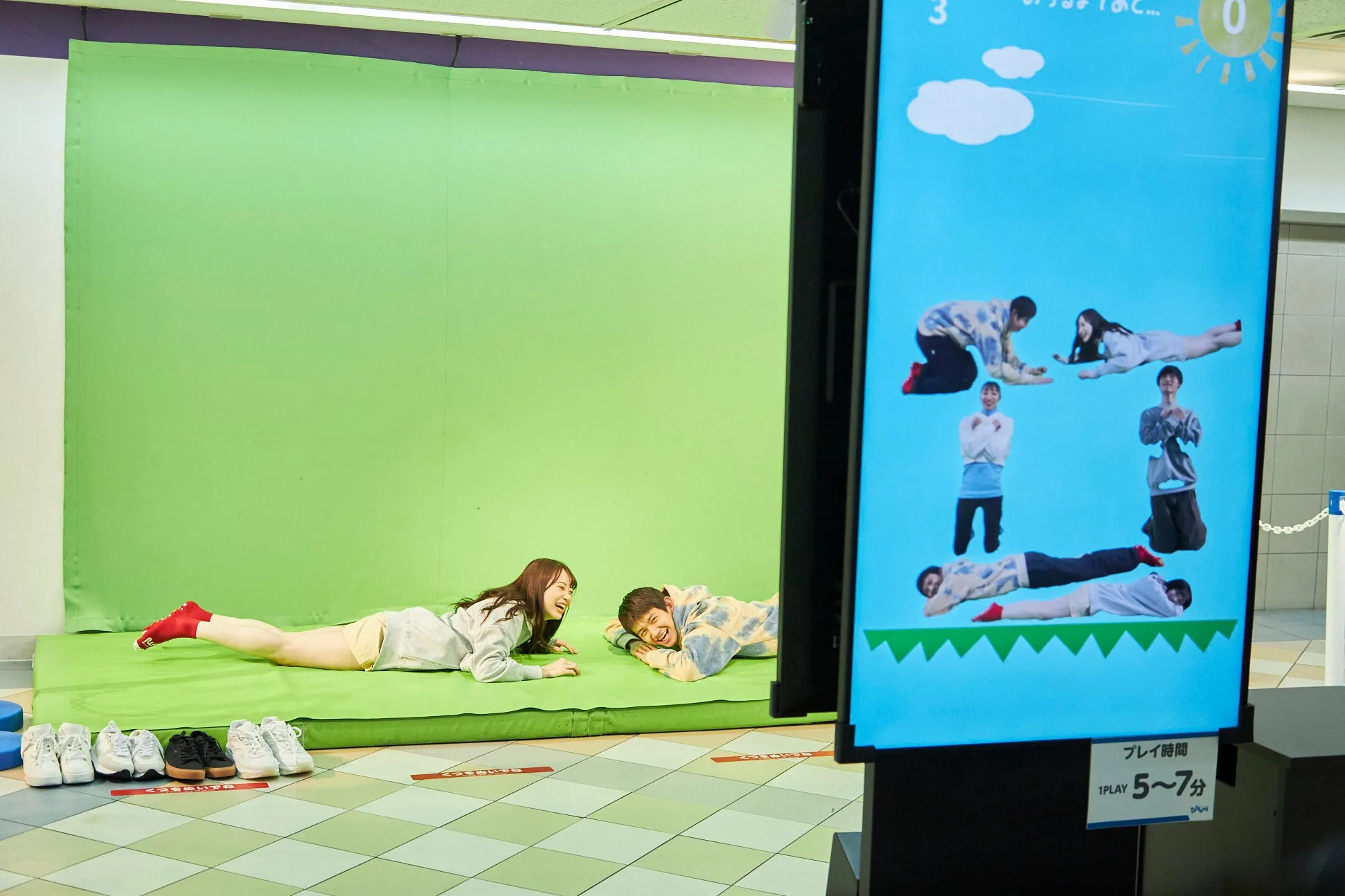 二人の若者が床に寝そべり、モニターに表示されたゲームを楽しんでいる様子を捉えた写真です。モニターには、同様に床に寝そべった人々の画像が表示されています。二人の周りには、たくさんの靴が並べられています。