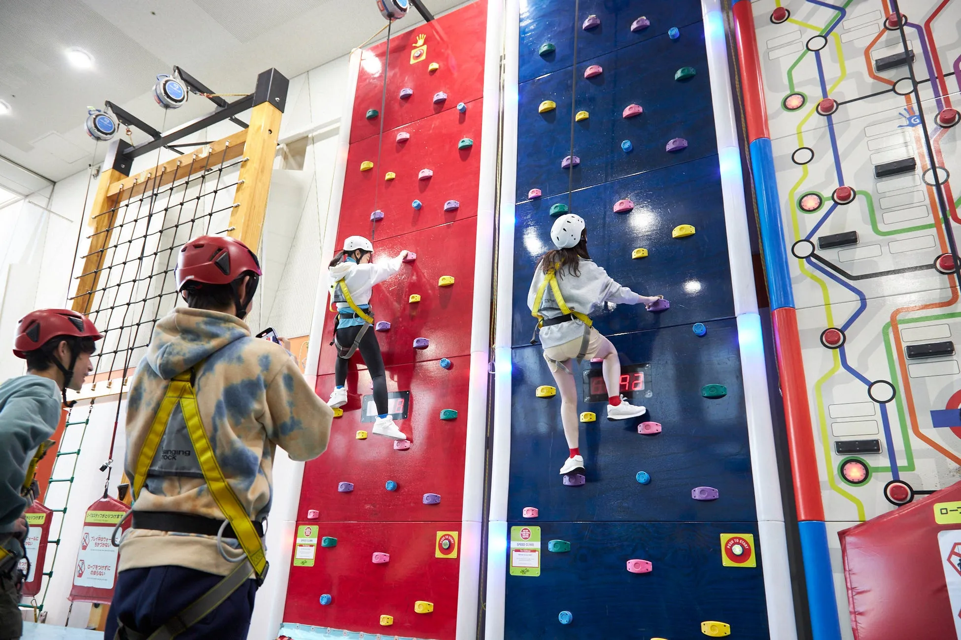 クライミングウォールでクライミングをする人々の様子。赤と青の壁があり、それぞれに人が登っています。観客もいます。