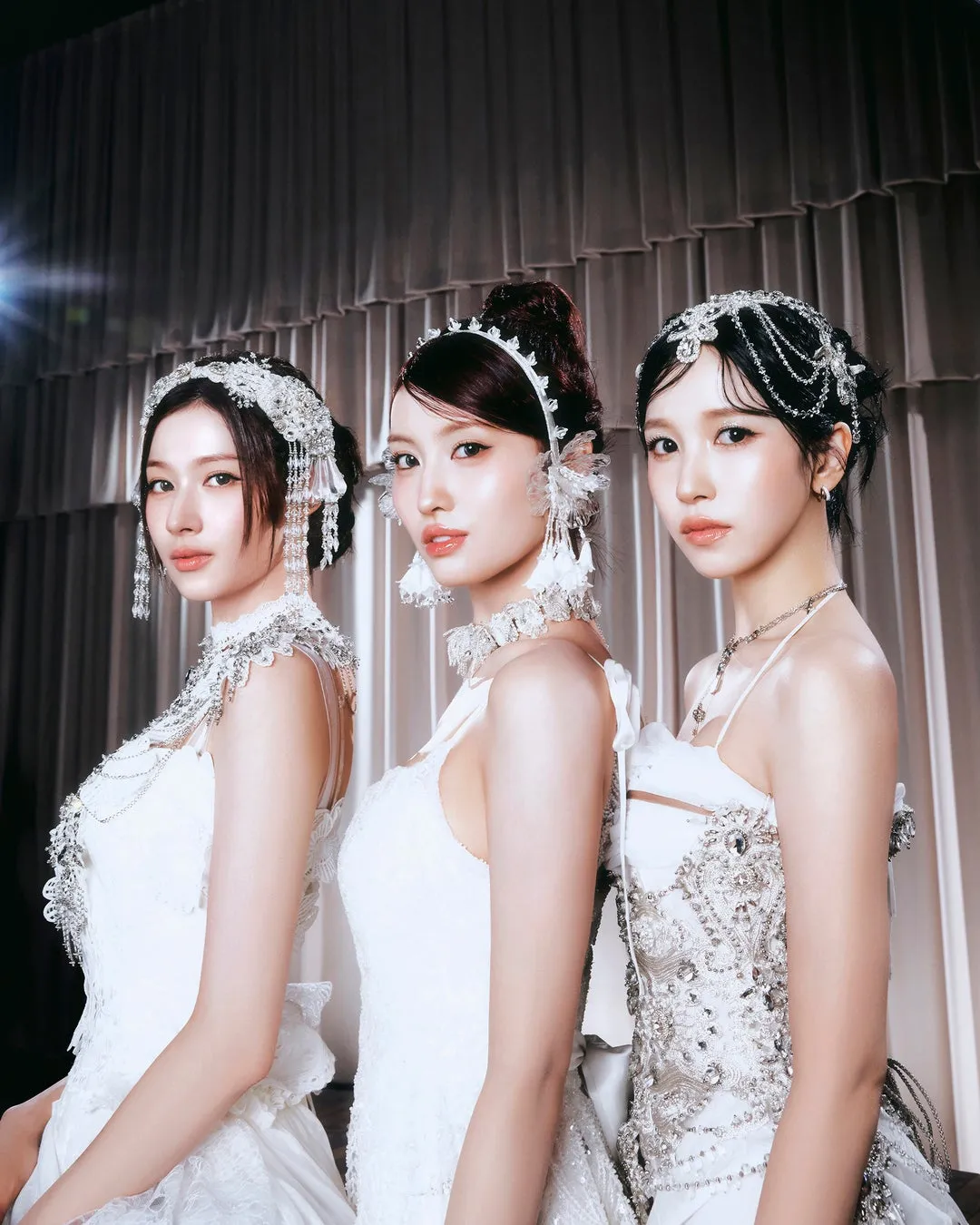 3人の女性が白いドレスを着てポーズをとっている写真。女性たちはそれぞれ、精巧な装飾が施された白いヘッドバンドとジュエリーを身につけている。背景はカーテンで、照明が雰囲気を作り出している。