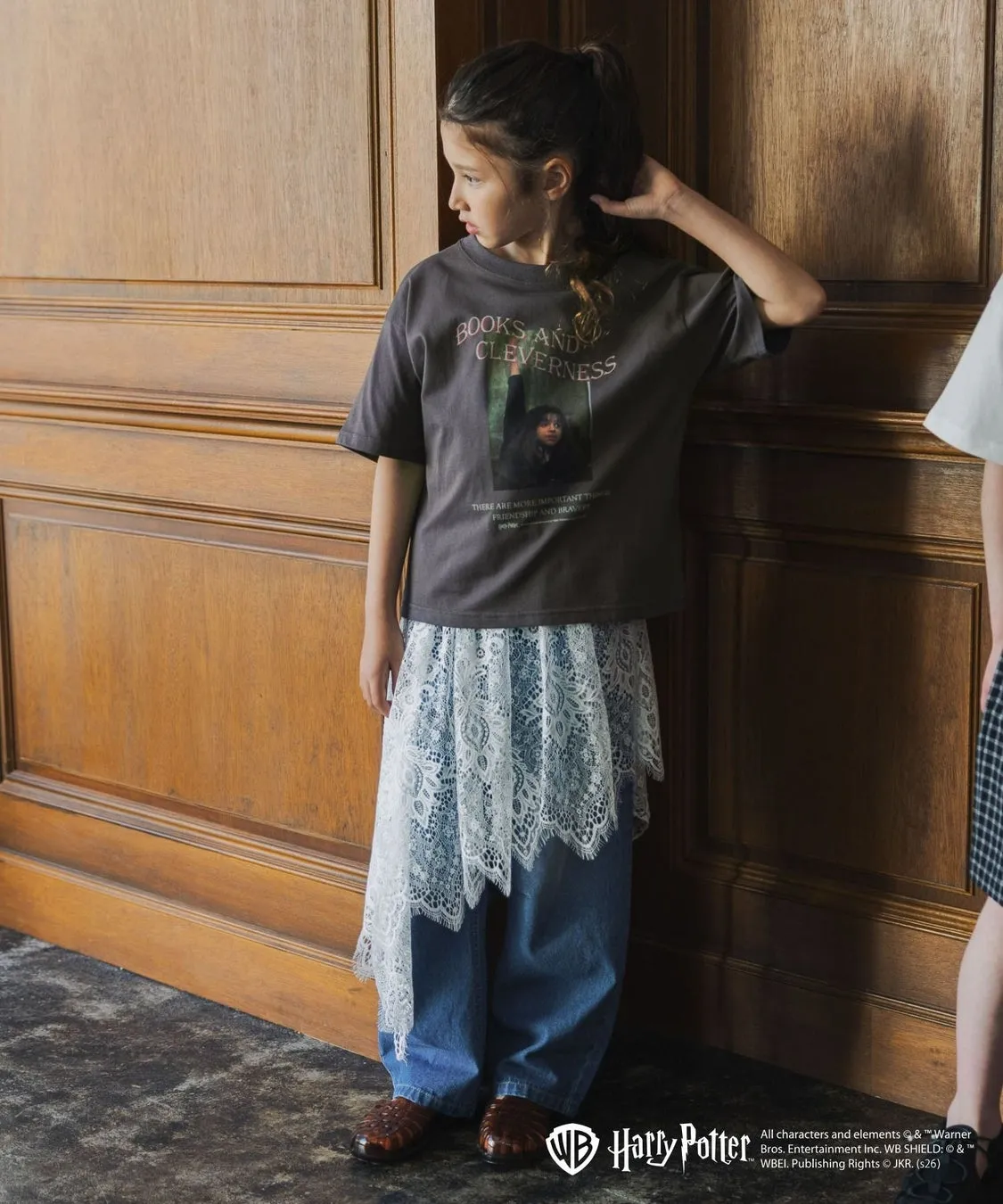 木製の壁の前に立っている、Tシャツとジーンズを着た若い女の子。Tシャツには「BOOKS AND CLEVERNESS」というテキストと、ハリーポッターのキャラクターの画像がプリントされている。彼女は茶色の革靴を履いている。