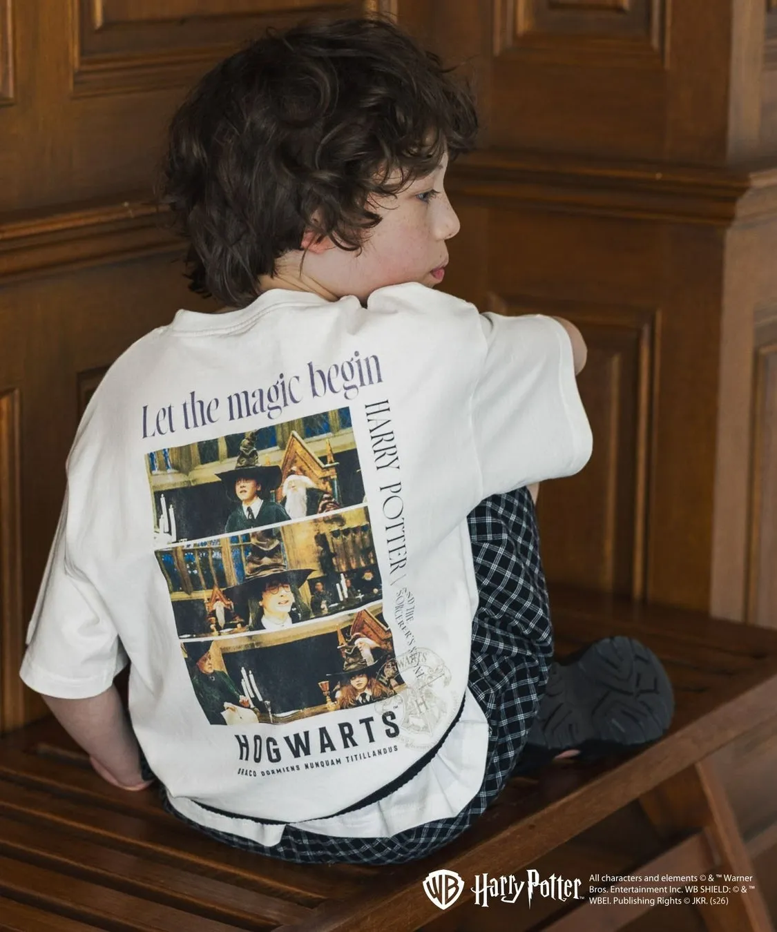 子供がハリーポッターのTシャツを着てベンチに座っている写真です。Tシャツには「Let the magic begin」と「HOGWARTS」の文字と映画のシーンがプリントされています。