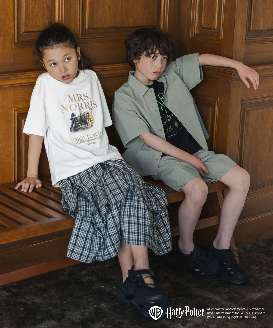 2人の子供がベンチに座っている写真。女の子はハリーポッターのMrs. NorrisのTシャツとチェック柄のスカートを着用し、男の子はグリーンのシャツとショートパンツを着用しています。背景は木製の壁です。