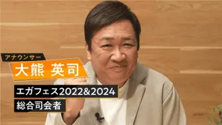 男性アナウンサーの顔のアップ。背景は薄い色の壁と木製のテーブルで、アナウンサーは笑顔でカメラを見ている。