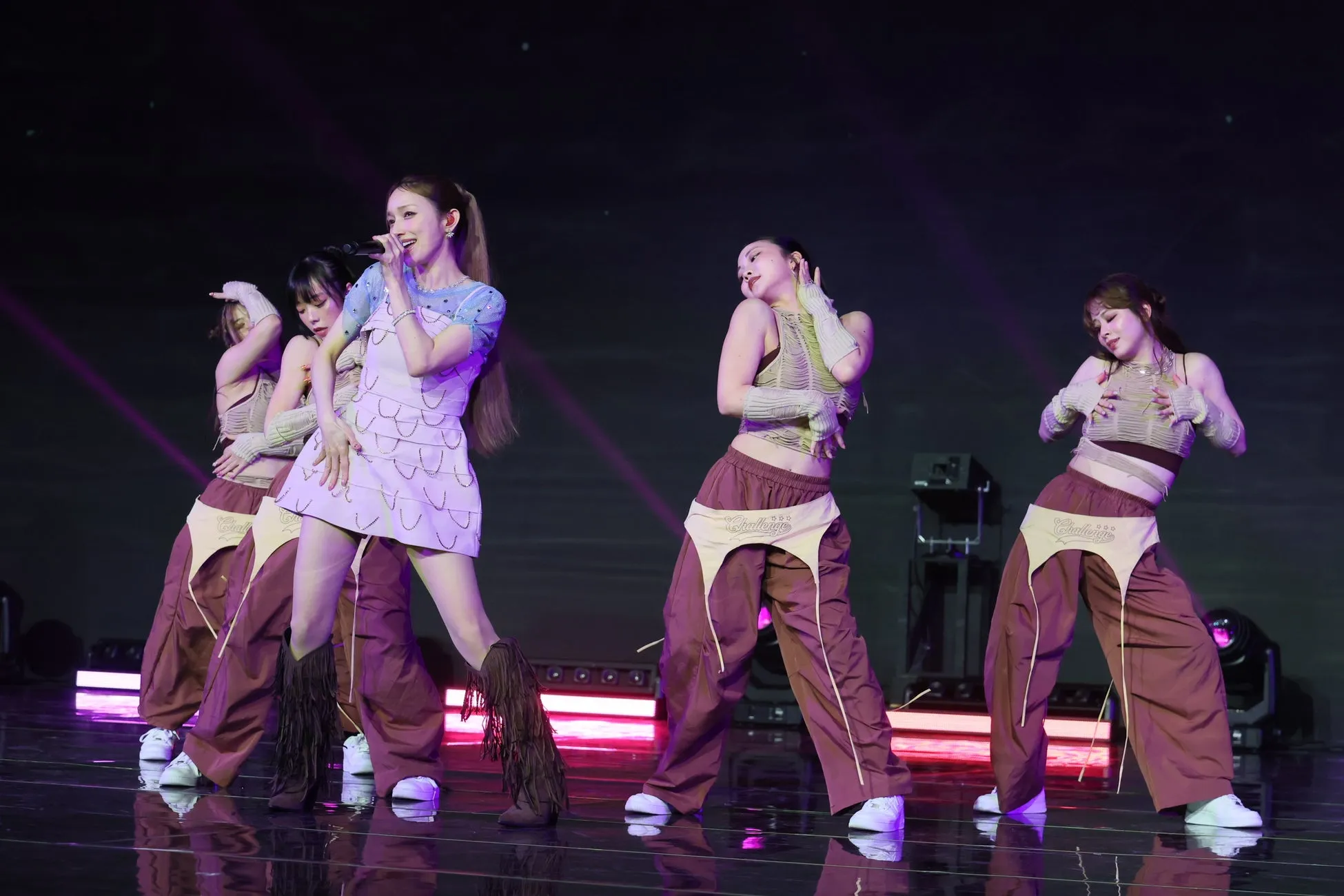 ステージで歌い踊る女性アイドルグループの画像。中央の女性がマイクを持ち歌い、他のメンバーはダンスをしている。