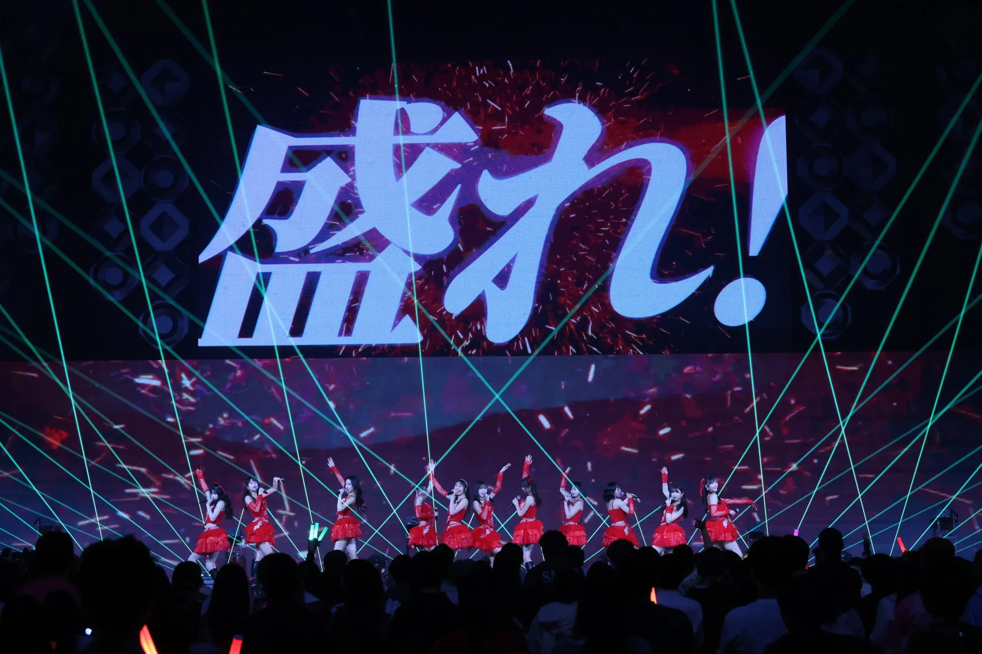 ステージでパフォーマンスをするアイドルグループの写真。背景には「盛り上がれ！」という文字が映し出されている。