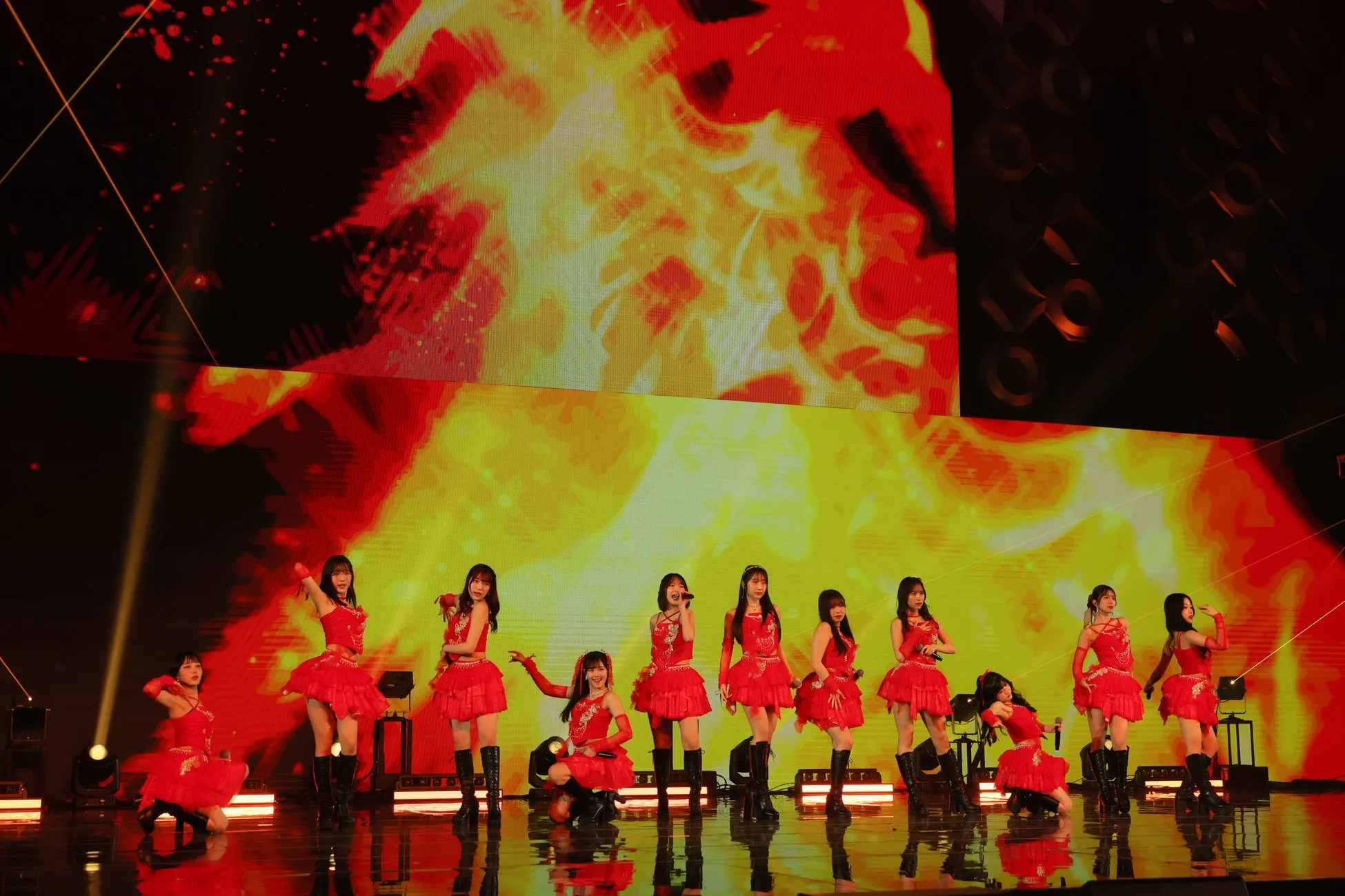 ステージでパフォーマンスをする女性アイドルグループの画像。メンバーは赤い衣装を着ており、ステージの背景は炎のようなデザイン。