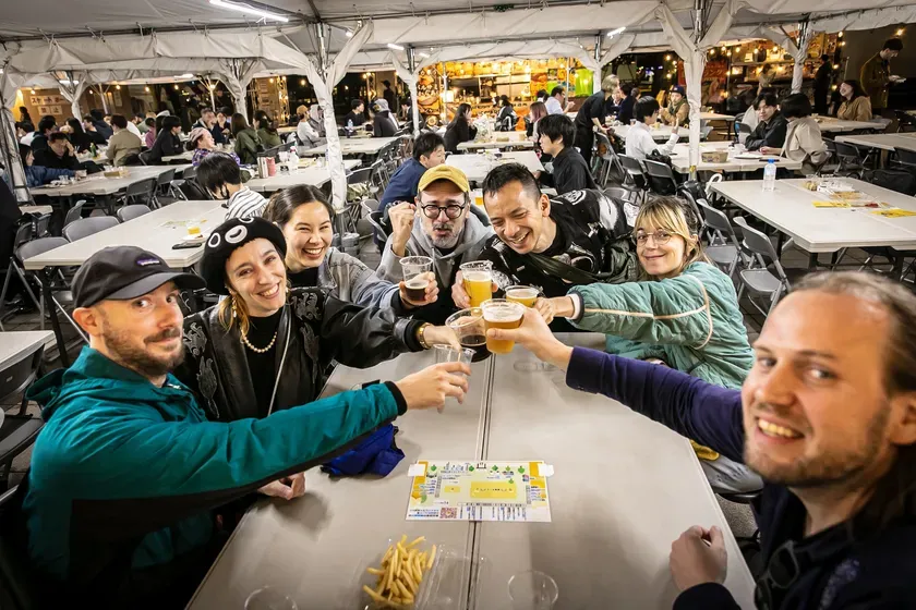 ビールを手に乾杯するグループの人々。イベント会場と思われる場所で、テーブルにはフライドポテトが置かれている。