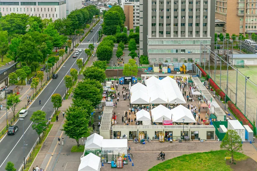 都市の鳥瞰図で、道路、建物、緑地、イベント会場が写っています。道路には車が通り、イベント会場には多くの人が集まっています。