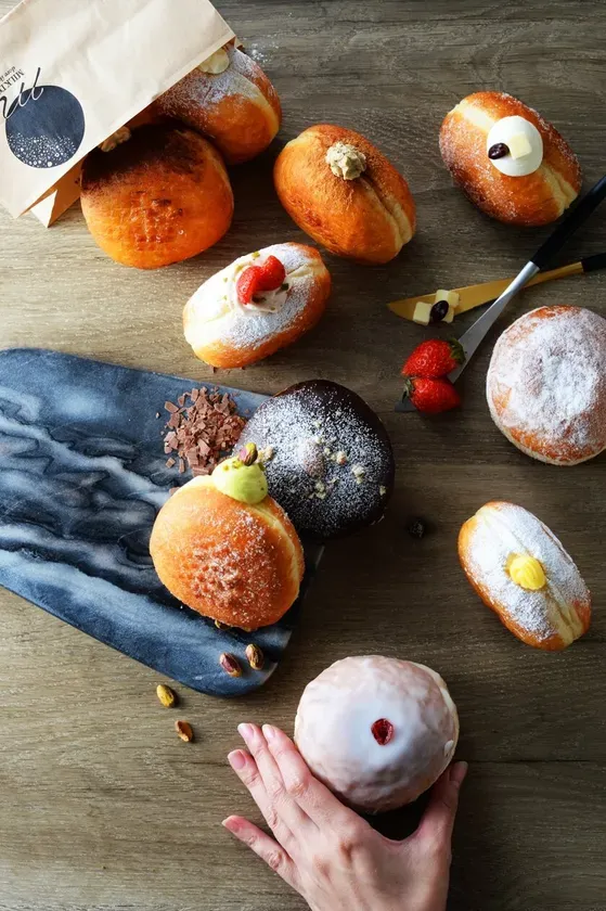 様々な種類のドーナツがテーブルに並べられた写真。ドーナツは、クリーム、ストロベリー、チョコレートなどでトッピングされており、美味しそうに見えます。手でドーナツを持っている様子も写っています。