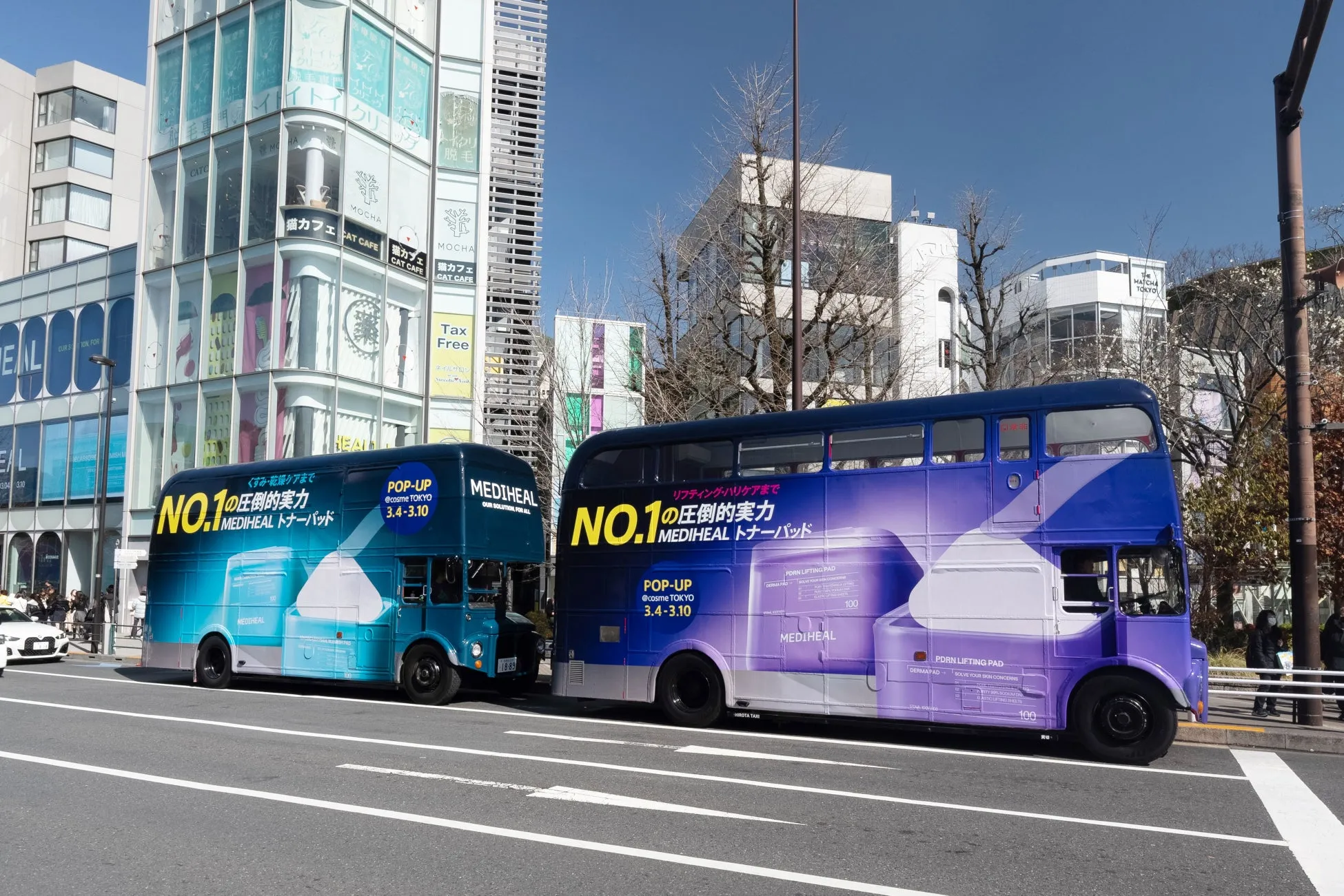 2台の二階建てバスが道路に停車している写真。バスにはMEDIHEALの広告が大きく表示されており、コスメ関連のイベント告知や製品情報が記載されている。