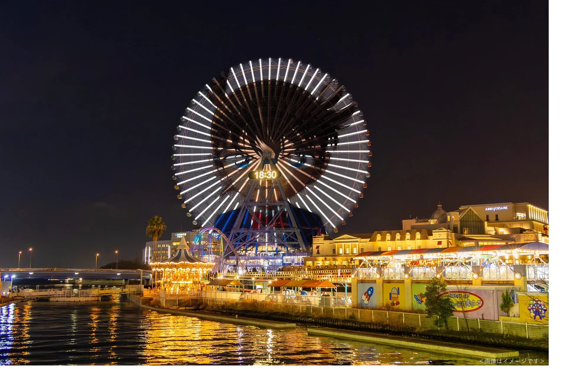 夜の観覧車と周辺の建物の写真。観覧車には18:30の時刻が表示され、ライトアップされている。水面に光が反射し、遊園地のような雰囲気。
