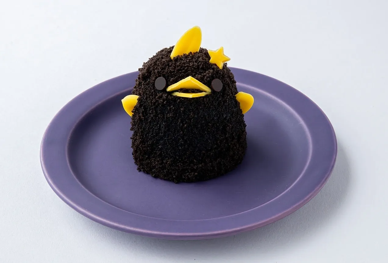 紫色の皿の上に、鳥の形をしたチョコレートケーキが置かれています。ケーキは、黒いクリームで覆われており、黄色い飾りで鳥の顔と翼が表現されています。