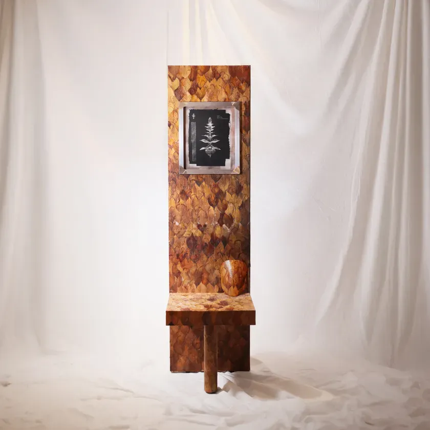 茶色を基調とした、葉の模様が施された背もたれと、その下に小さな台座がついた椅子のようなオブジェ。正面には植物の絵が飾られており、台座には壺のようなものが置かれている。背景は白い布で覆われている。