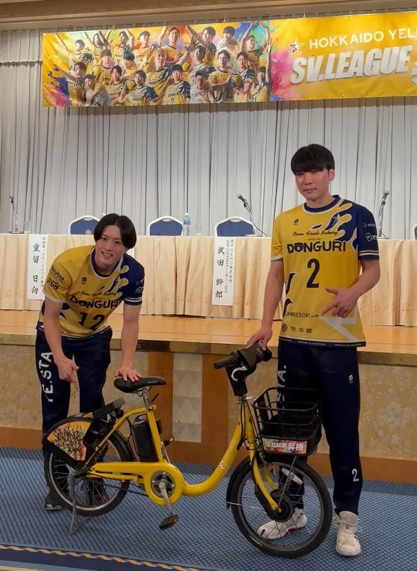 2人の男性が自転車の横に立っている写真です。2人は黄色のユニフォームを着ています。背景には、SV.LEAGUEのバナーとテーブルがあります。