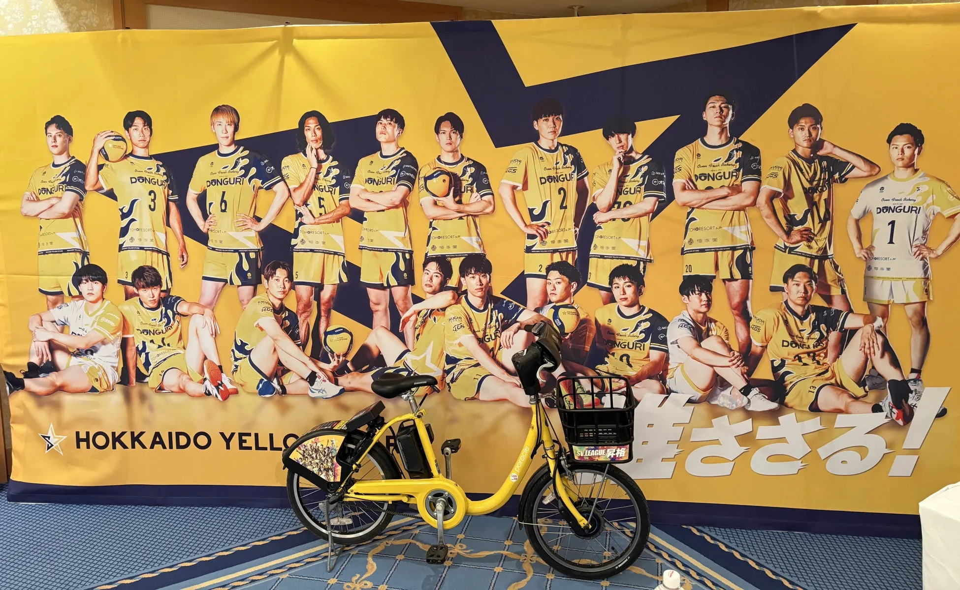 バレーボールチームの選手たちと自転車が写った写真。背景にはチームのロゴとスローガンが描かれている。