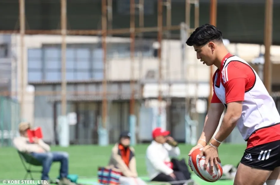 ラグビーボールを構える男性が写っている写真です。周りには、座って本を読む人や、飲み物を飲んでいる人など、数人の人がいます。