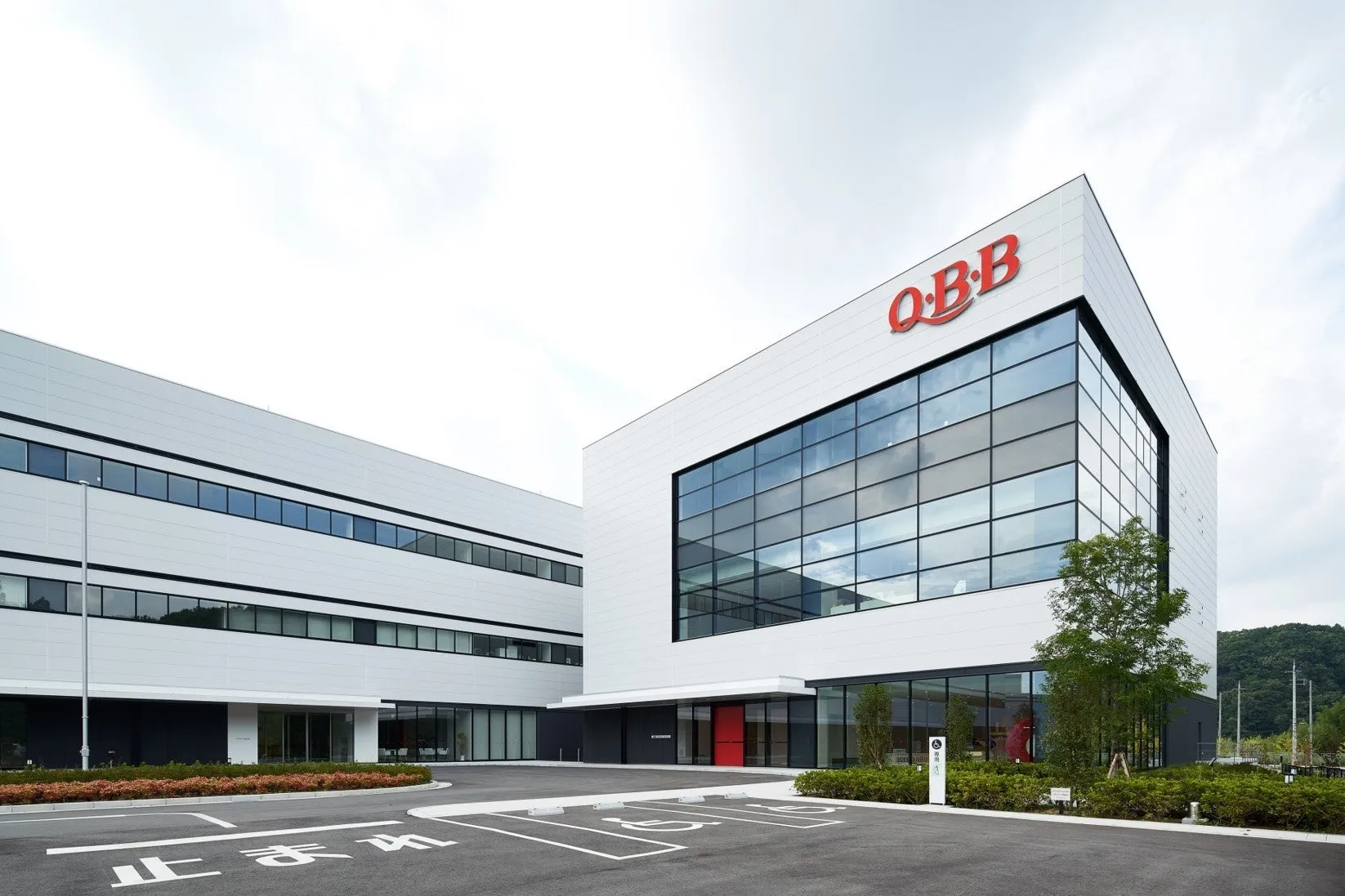 白いモダンな建物。建物の正面には「O・B・B」のロゴがあり、大きな窓がある。建物の前には駐車場と緑地があり、空は曇っている。