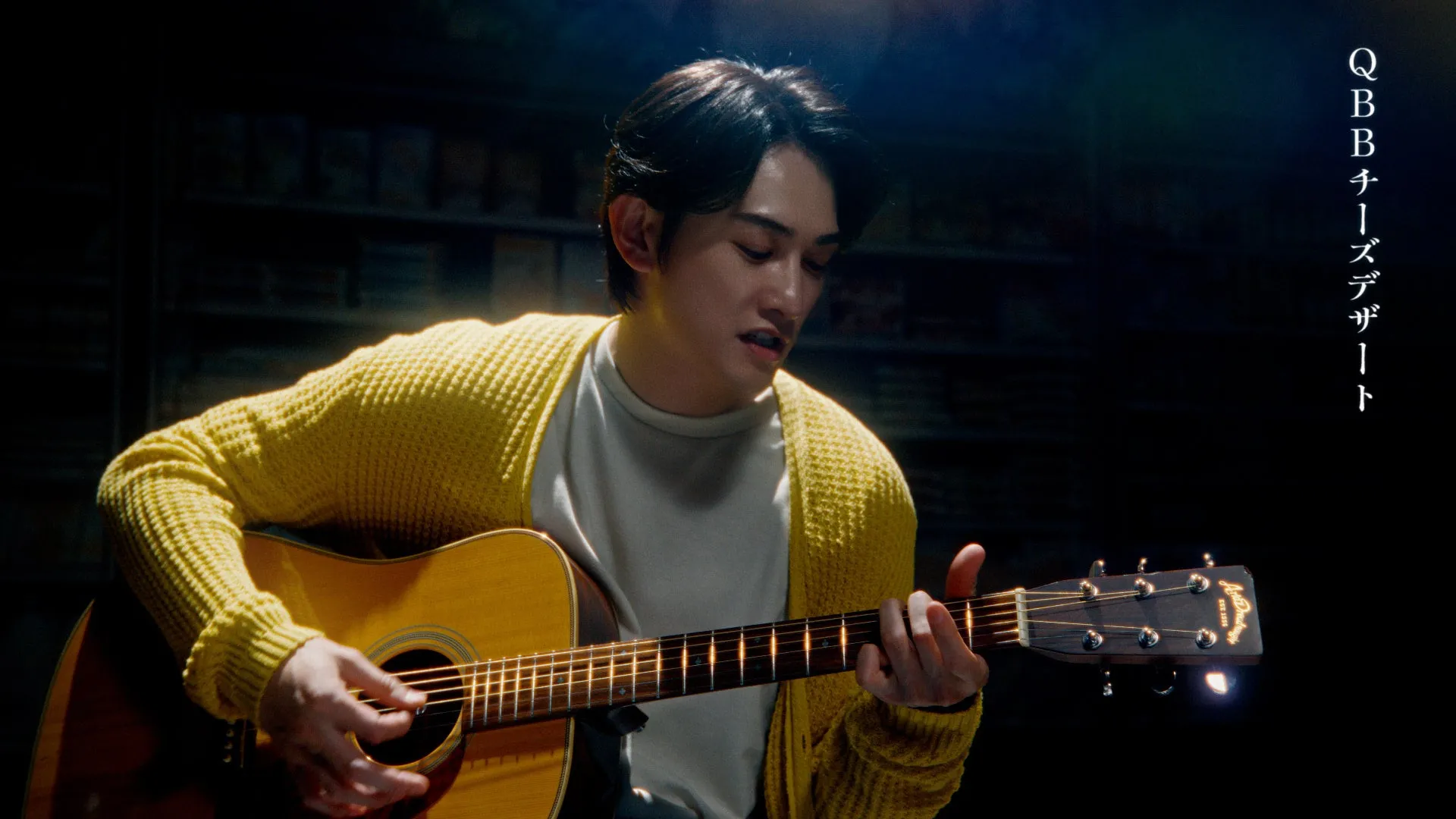 男性がアコースティックギターを演奏している様子を捉えた画像。彼は黄色のカーディガンを着ており、集中した表情でギターを弾いている。背景は暗く、照明が彼とギターを照らしている。