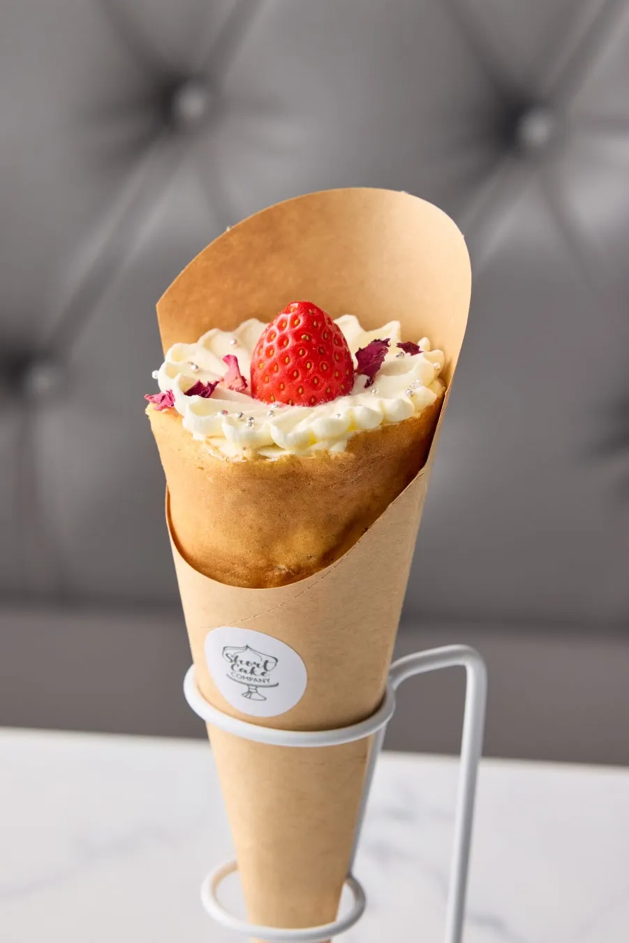 ストロベリーとクリームがトッピングされた、茶色の紙のコーンに入ったデザートのクローズアップ写真。デザートは、白いスタンドに置かれています。