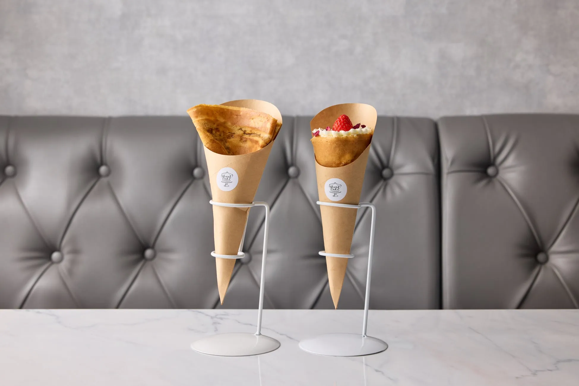 2つのコーンに入ったデザートがテーブルの上に置かれています。デザートは、紙製のコーンに入っており、スタンドに立てられています。背景には灰色の革張りの壁があります。