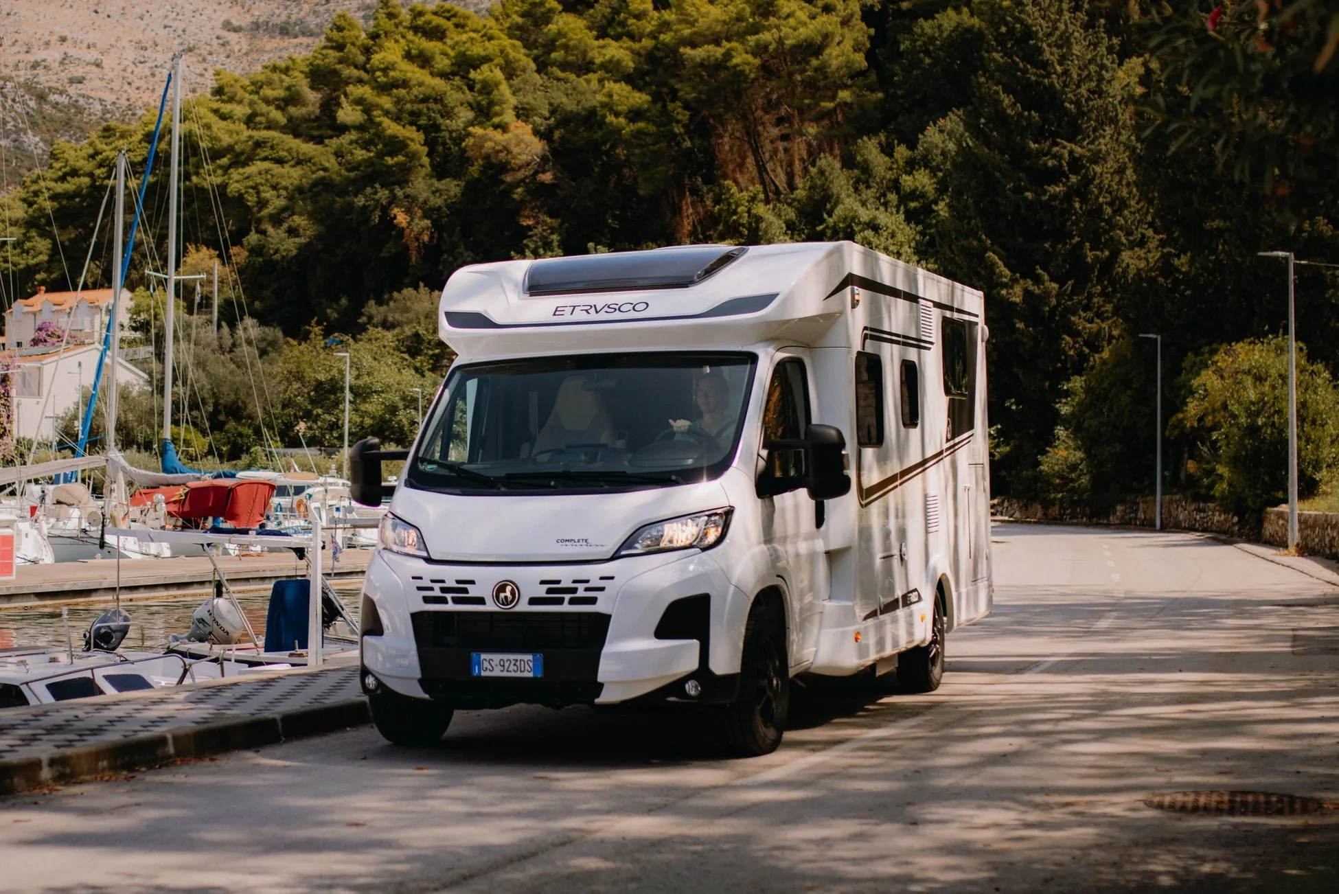 白いキャンピングカーがヨットハーバーに面した道を走行している様子