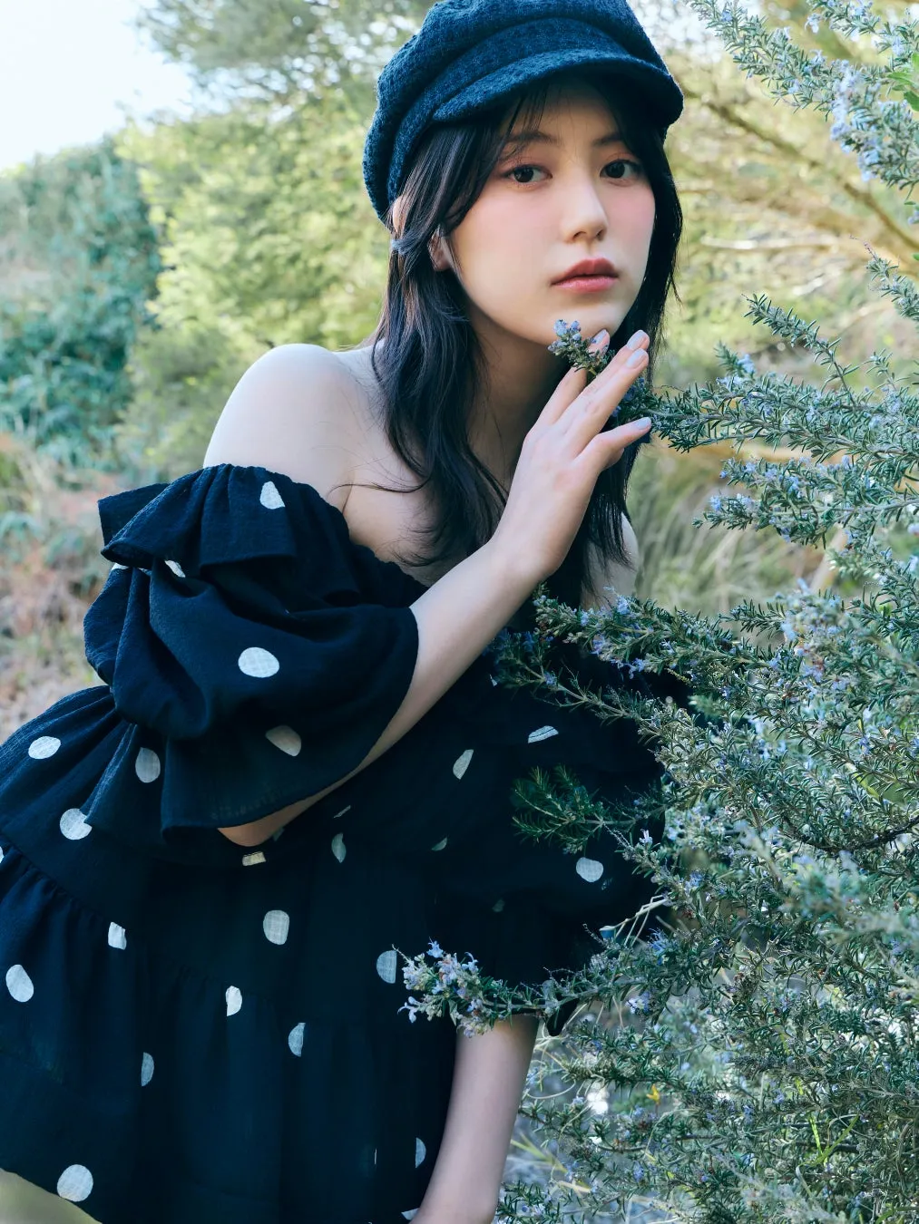 女性が、黒い帽子と水玉模様のオフショルダーのドレスを着て、屋外の植物に触れている写真です。