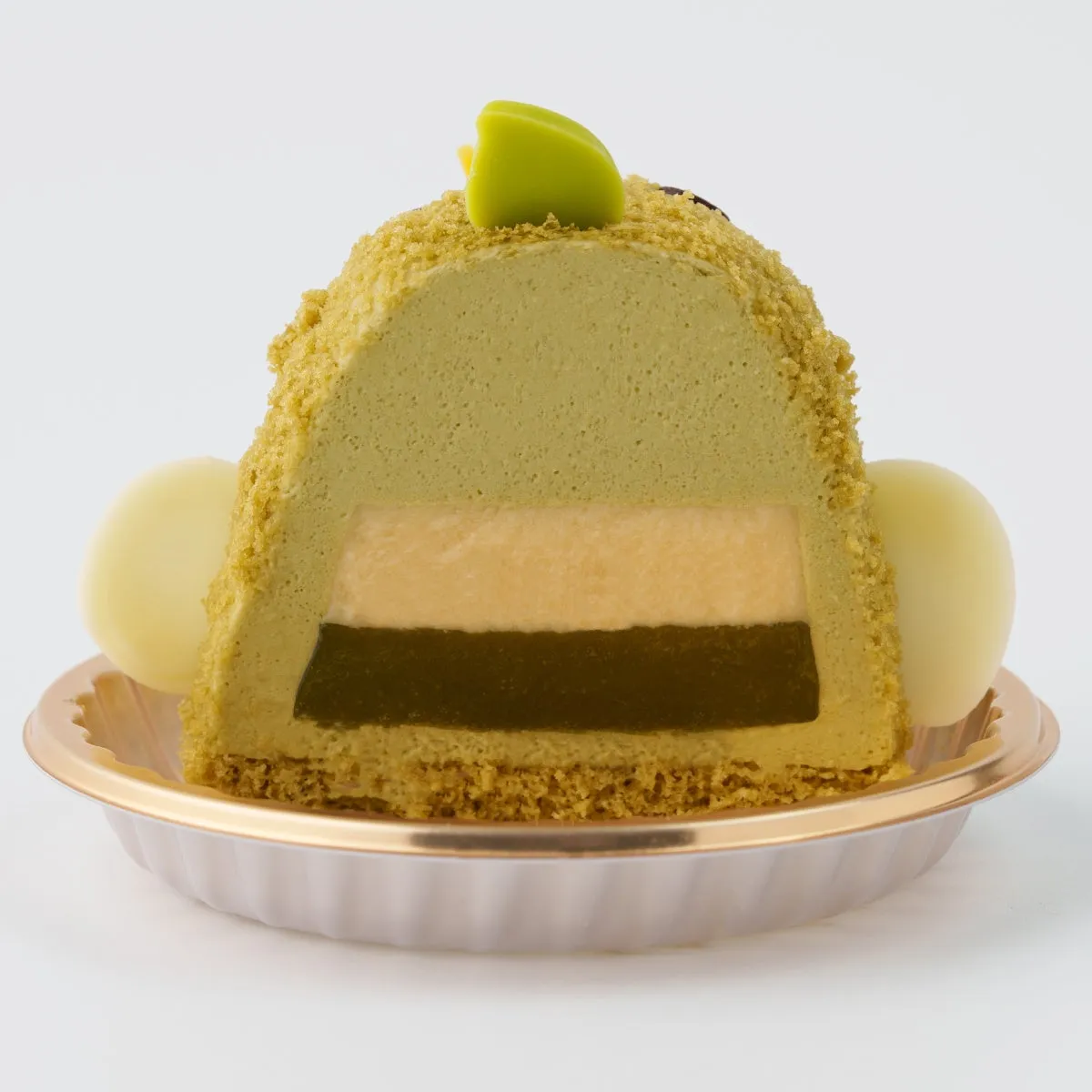 抹茶ケーキの断面図。抹茶ムース、オレンジ色の層、濃い緑色の層が特徴的で、白いチョコレートボールと緑色の飾りで飾られています。ケーキは金色の縁が付いた白い皿に載っています。