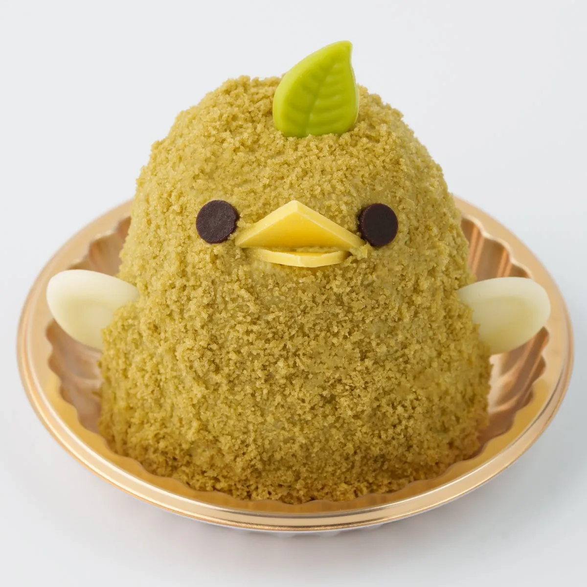鳥の形をしたケーキが、金色の皿に乗っている写真です。ケーキは緑色のクリームで覆われており、チョコレートの目と黄色いチョコレートのくちばし、葉の飾りが施されています。