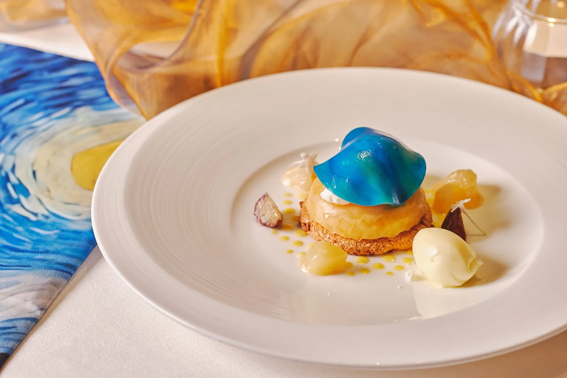 白い皿の上に、青いゼリー状のものが乗ったデザートが盛り付けられています。デザートは、クッキーのような土台の上に、オレンジ色の層が重なり、その上に青いゼリー状のものが乗っています。デザートの周りには、小さな果物やソース、アイスクリームが添えられています。背景には、テーブルクロスと、絵画風の布が写っています。