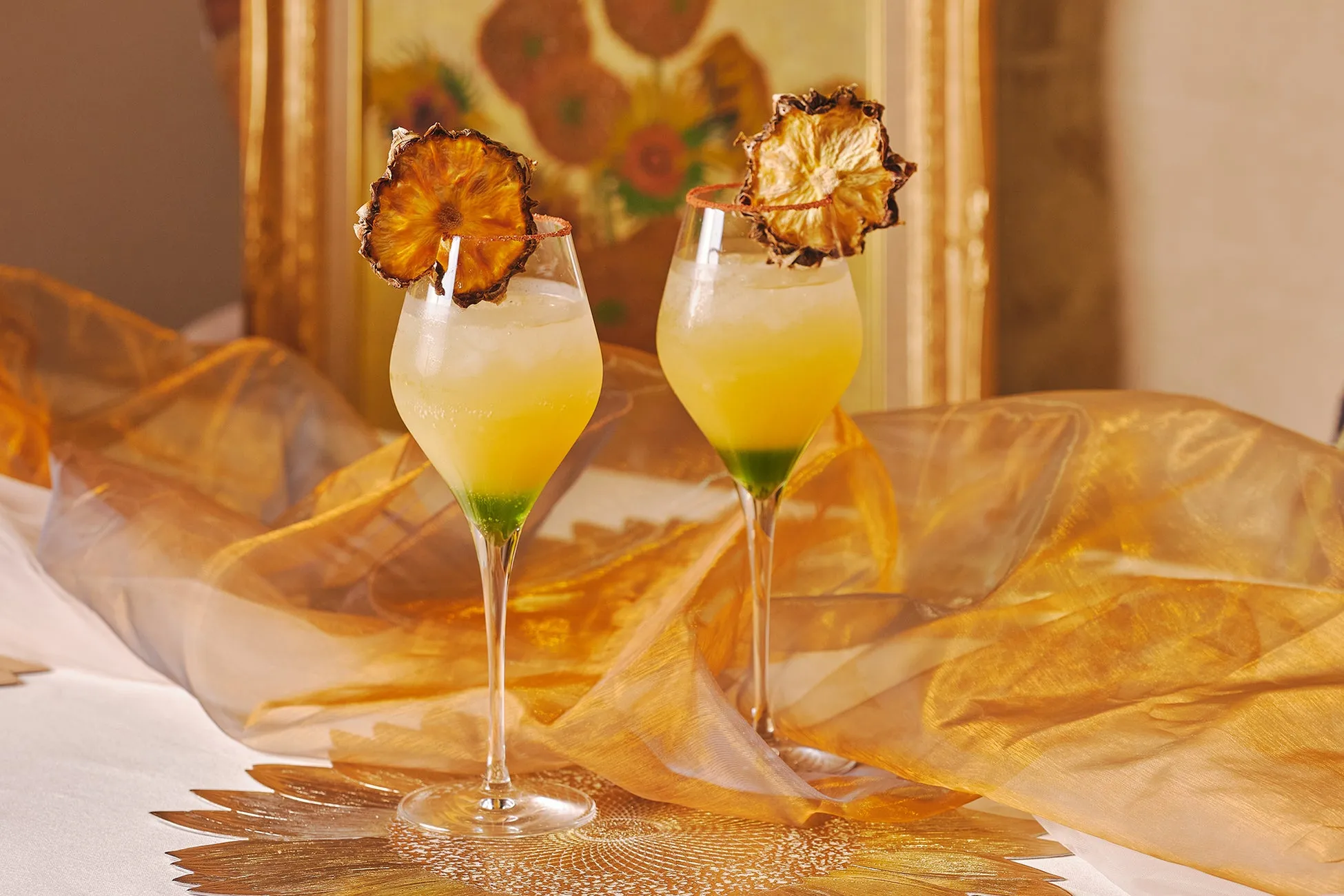 2つのグラスに入った黄色いカクテル。グラスの縁には赤い砂糖がまぶされ、パイナップルのドライフルーツが添えられている。背景には絵画と金色の布がある。