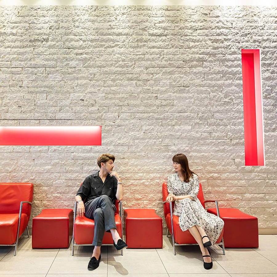 二人の男女が赤い椅子に座って話している写真。背景は石壁で、赤い照明がアクセントになっている。