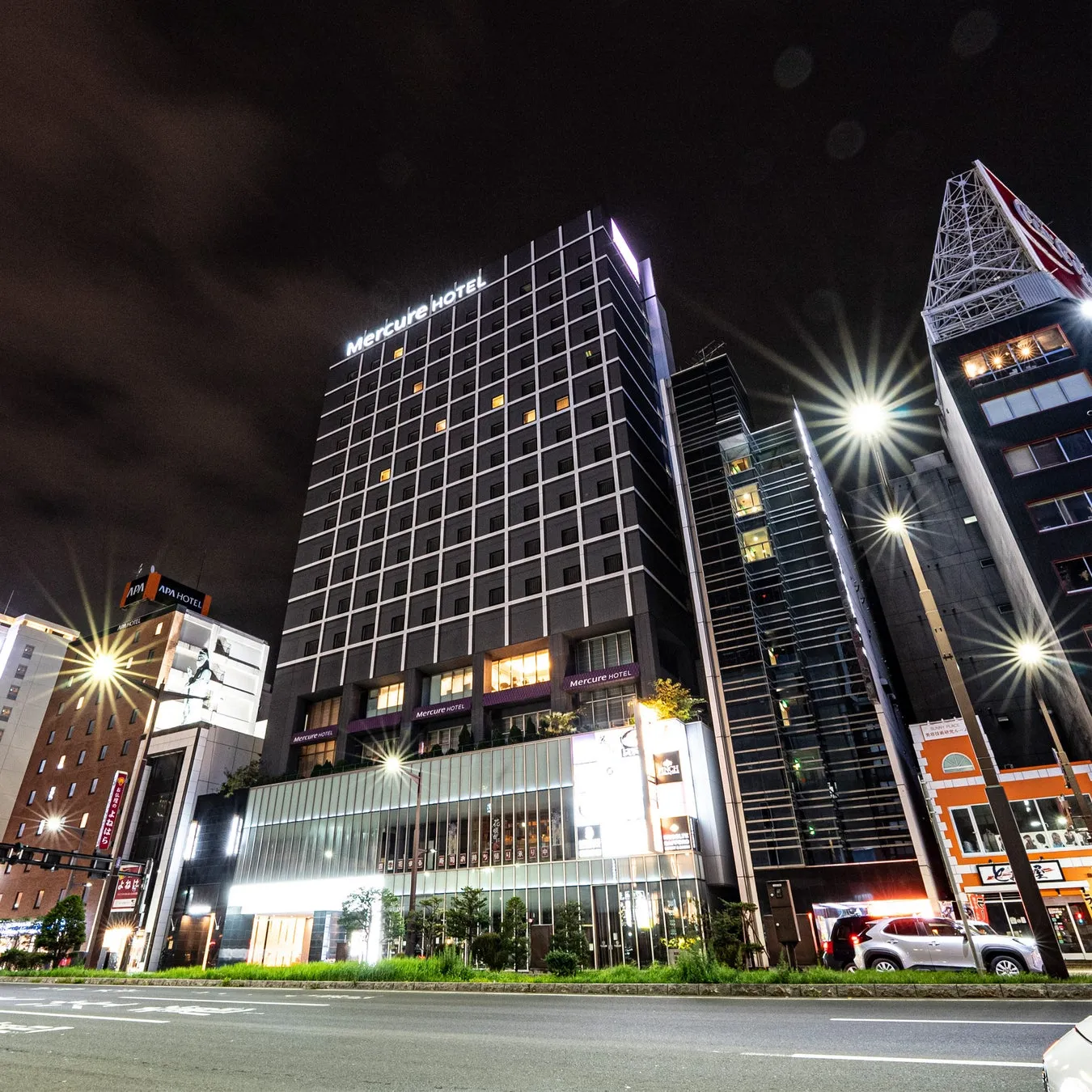 夜の街並みの写真。Mercure HotelとAPA HOTELの看板が目立つ。道路には街灯があり、車が走っている。