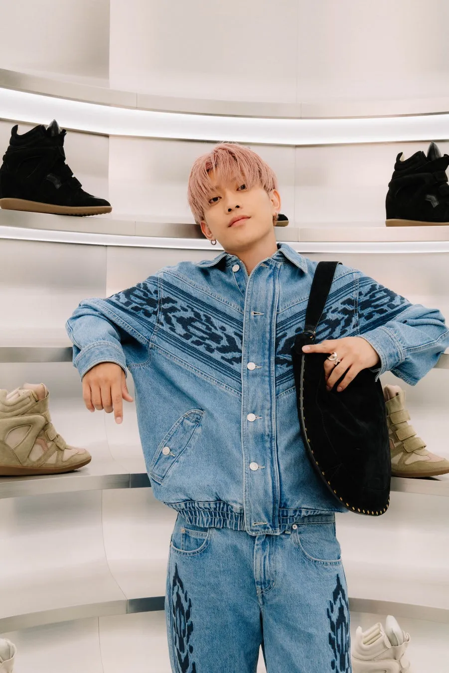 ピンク色の髪の男性が、ジーンズのジャケットとパンツを着て、黒いバッグを持っています。背景には靴がディスプレイされています。