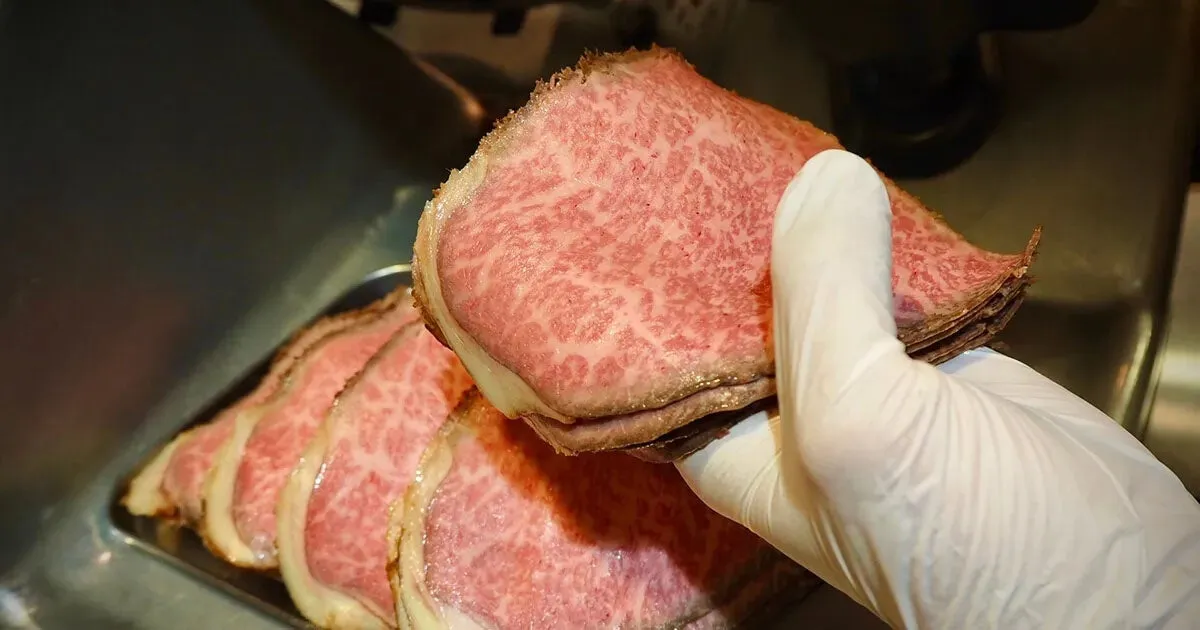 手袋をした手が、ローストビーフの切り身をいくつか持っている様子を捉えた写真です。肉は非常に高品質で、美しいマーブリングが特徴です。