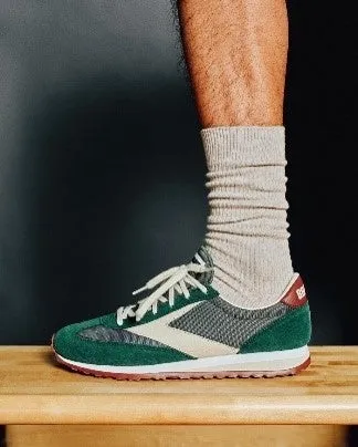 緑色のスニーカーを履いた足のクローズアップ写真。スニーカーは、白いレース、茶色のソール、白い「B」のロゴが入った緑色のスエードで作られています。足には、ベージュの靴下も履いています。