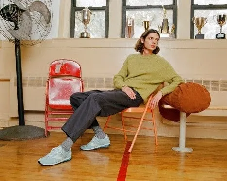 男性が椅子に座って振り返っている写真。彼は緑色のセーターと黒いズボン、水色の靴を履いています。背景には窓があり、トロフィーが飾られています。