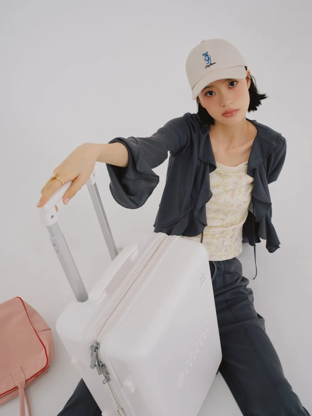 白いスーツケースとピンクのバッグを持った女性が、ライトベージュのキャップを被り、カメラ目線で座っている様子。