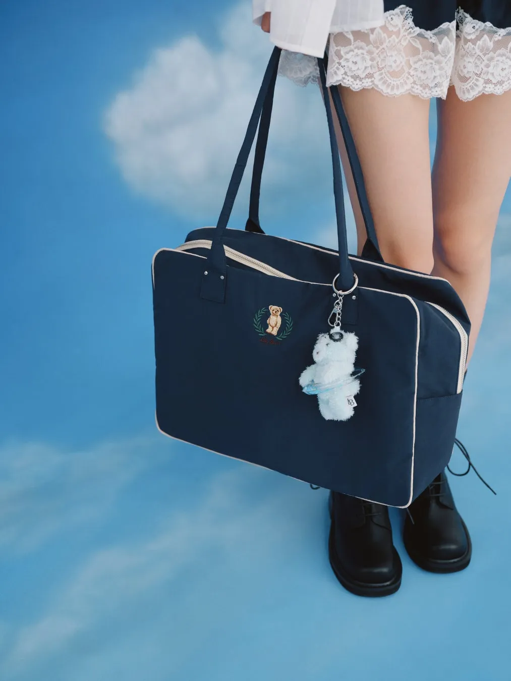 青空を背景に、ネイビーのバッグを持った女性の姿。バッグにはクマの刺繍とクマのキーホルダーが付いている。女性は白い服と黒い革靴を履いている。