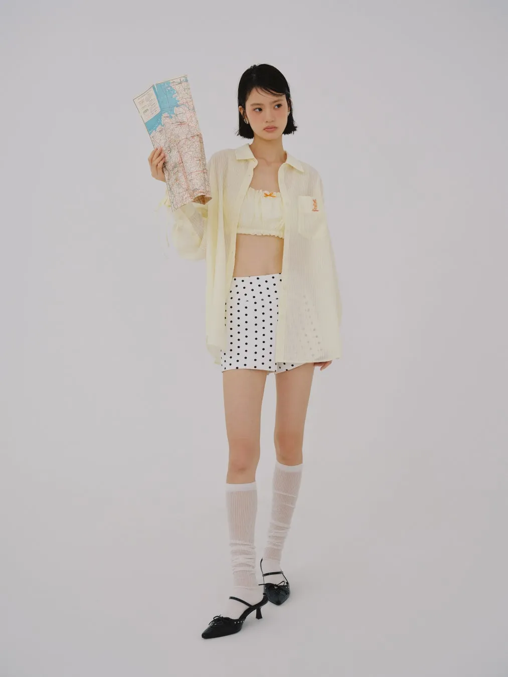 女性が地図を持ち、黄色いシャツ、白い水玉模様のスカート、白い靴下、黒い靴を履いて立っています。背景は白です。