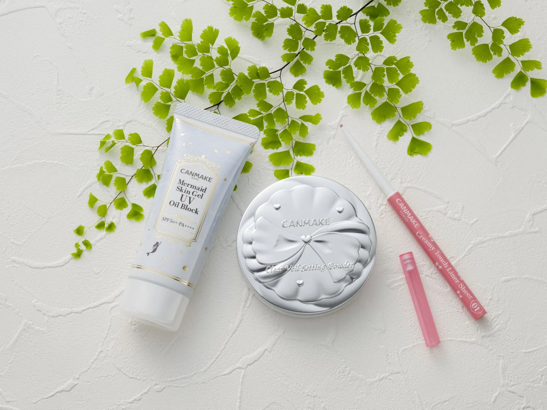 化粧品が置かれた写真。日焼け止め、パウダー、アイライナーが写っており、背景には葉が添えられている。