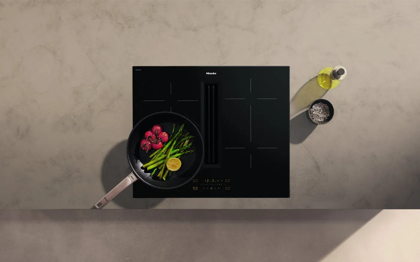 黒いコンロの上に、アスパラガス、トマト、レモンが入ったフライパンが置かれ、オリーブオイルと塩も添えられています。