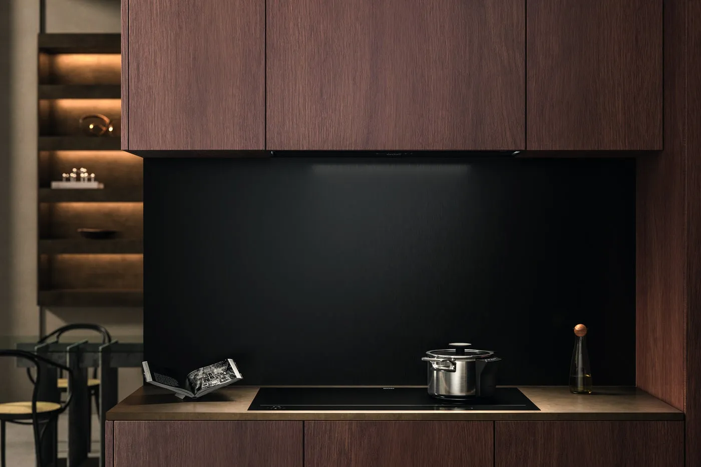 モダンなキッチンインテリアの写真。木製のキャビネットと黒い壁が特徴で、コンロ、鍋、開いた本、ガラス瓶がカウンターに置かれています。