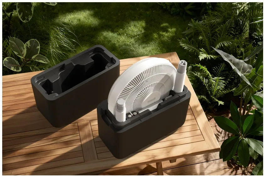 木製のテーブルの上に、ファンと収納ケースが置かれた画像です。ファンは折りたたまれた状態で収納ケースに収められており、もう一つの収納ケースは開いた状態です。背景には緑豊かな庭の様子が写っています。