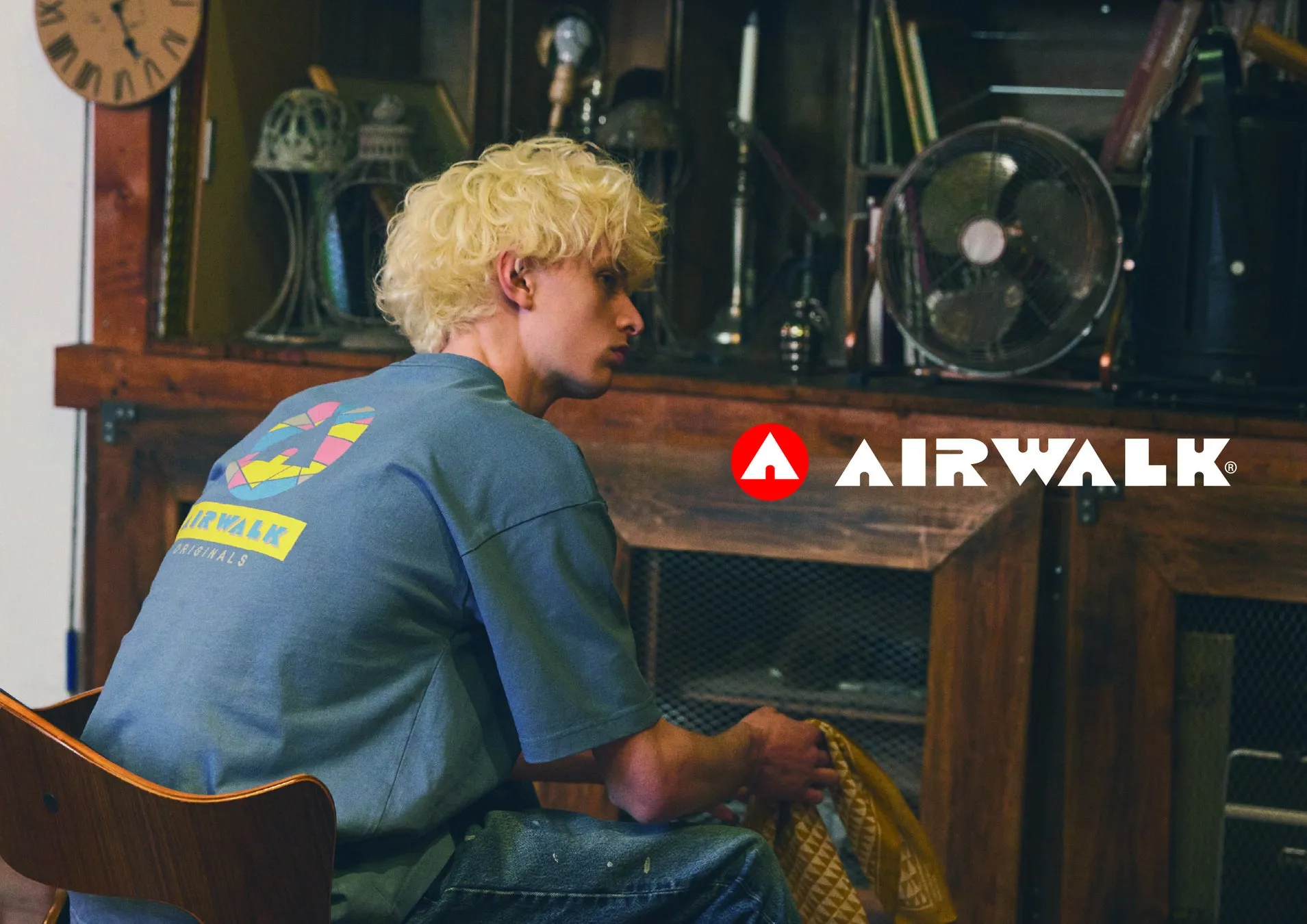 AIRWALKのTシャツを着て、椅子に座って何かを見ている金髪の男性。