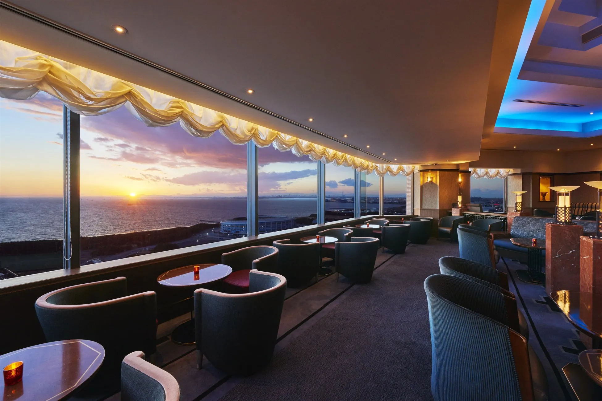 夕焼けの海を眺めることができる、高級感のあるバーのインテリアです。窓際にはテーブルと椅子が並び、落ち着いた雰囲気の中でくつろげます。照明も美しく、夕日の光が反射して、ロマンチックな空間を演出しています。