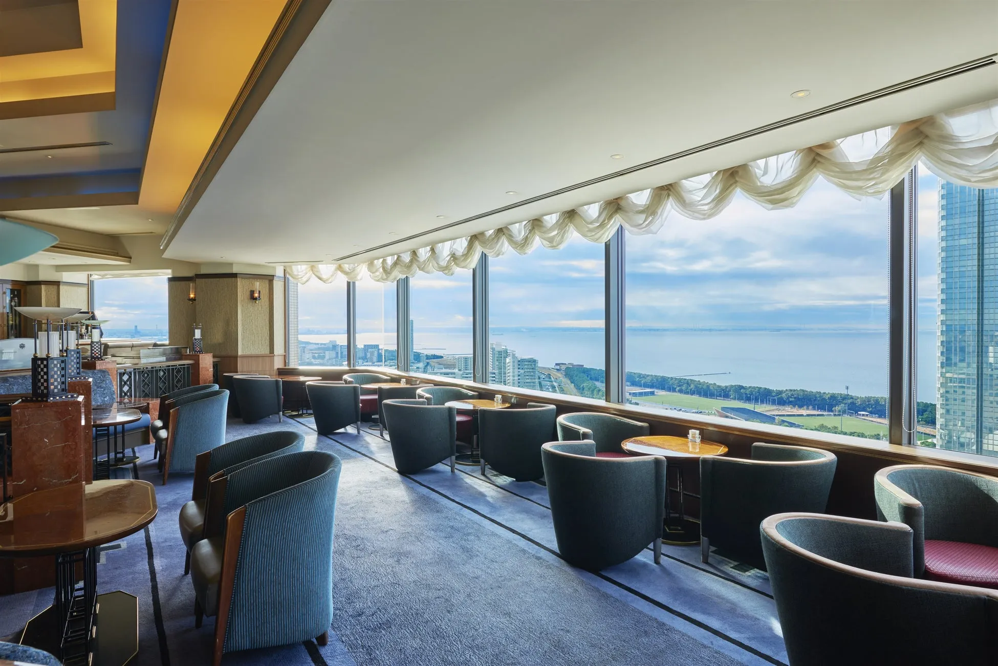 窓から海の見える、高級感のあるラウンジのインテリアです。丸い背もたれの椅子が並び、テーブルには飲み物が置かれています。窓からは、海と遠くの街並みが見えます。