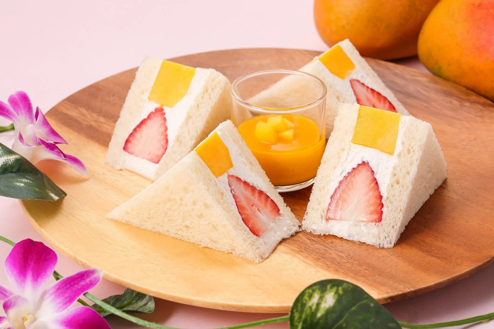 木製の皿に、イチゴとマンゴーが挟まれたサンドイッチとマンゴーソース、オレンジ、蘭の花が置かれた写真です。