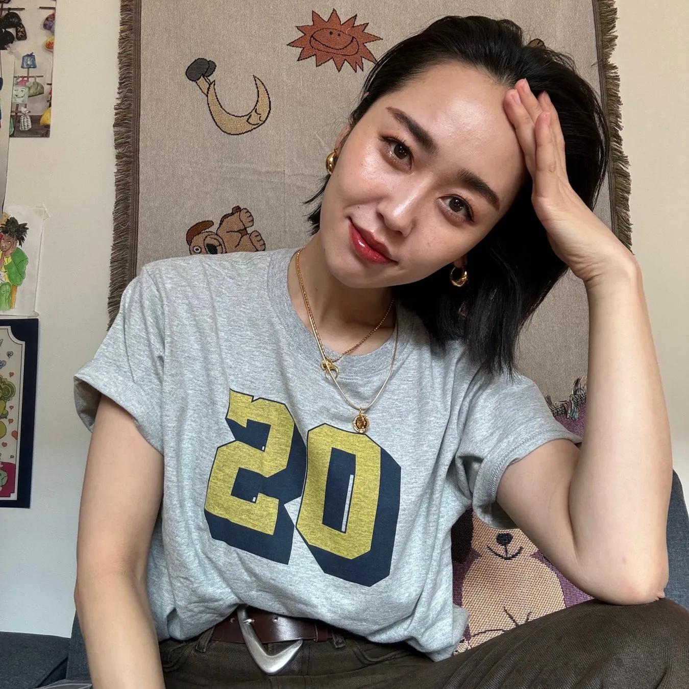 女性が正面を向き、手で頭を支えながら微笑んでいる写真。彼女はグレーのTシャツを着ており、数字の「20」がプリントされています。また、金色のネックレスとイヤリングを身に着けています。背景には、太陽や三日月、クマなどのイラストが描かれたタペストリーがあります。
