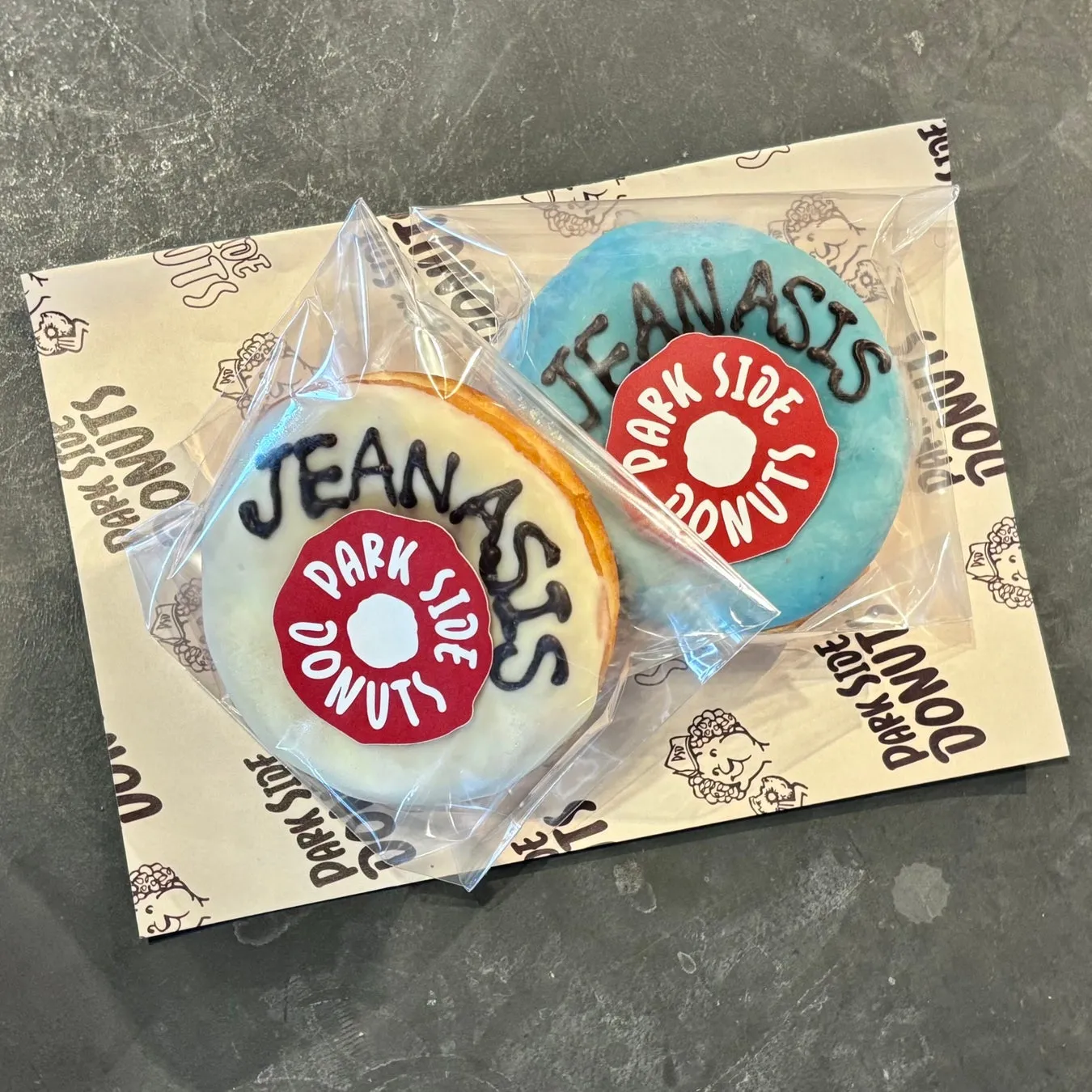 2つのドーナツが透明な袋に入っており、茶色の箱の上に置かれています。ドーナツには、白いアイシングの上に黒い文字で「JEANASIS」と書かれています。ドーナツの中央には「DARK SIDE DONUTS」と書かれた赤いロゴがあります。箱には「DARK SIDE DONUTS」の文字とイラストが印刷されています。
